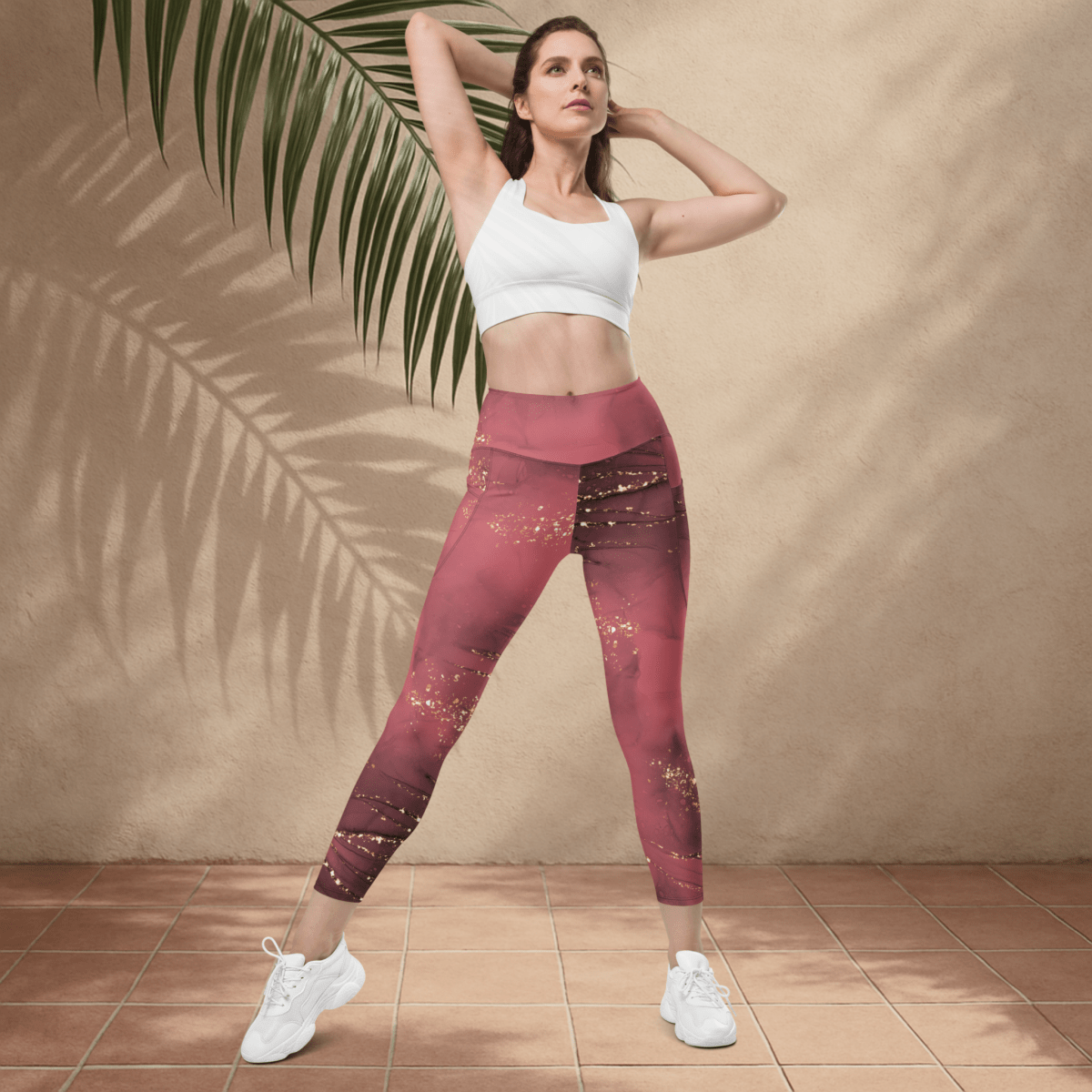 Woman wearing a white sports bra and dusty rose marble leggings standing against a beige wall with a palm leaf.