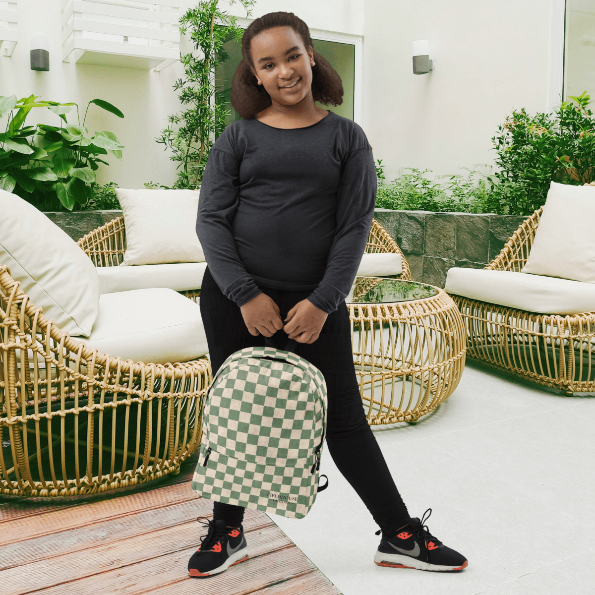 Person holding a green and white checkered backpack in an outdoor setting with wicker chairs and plants.