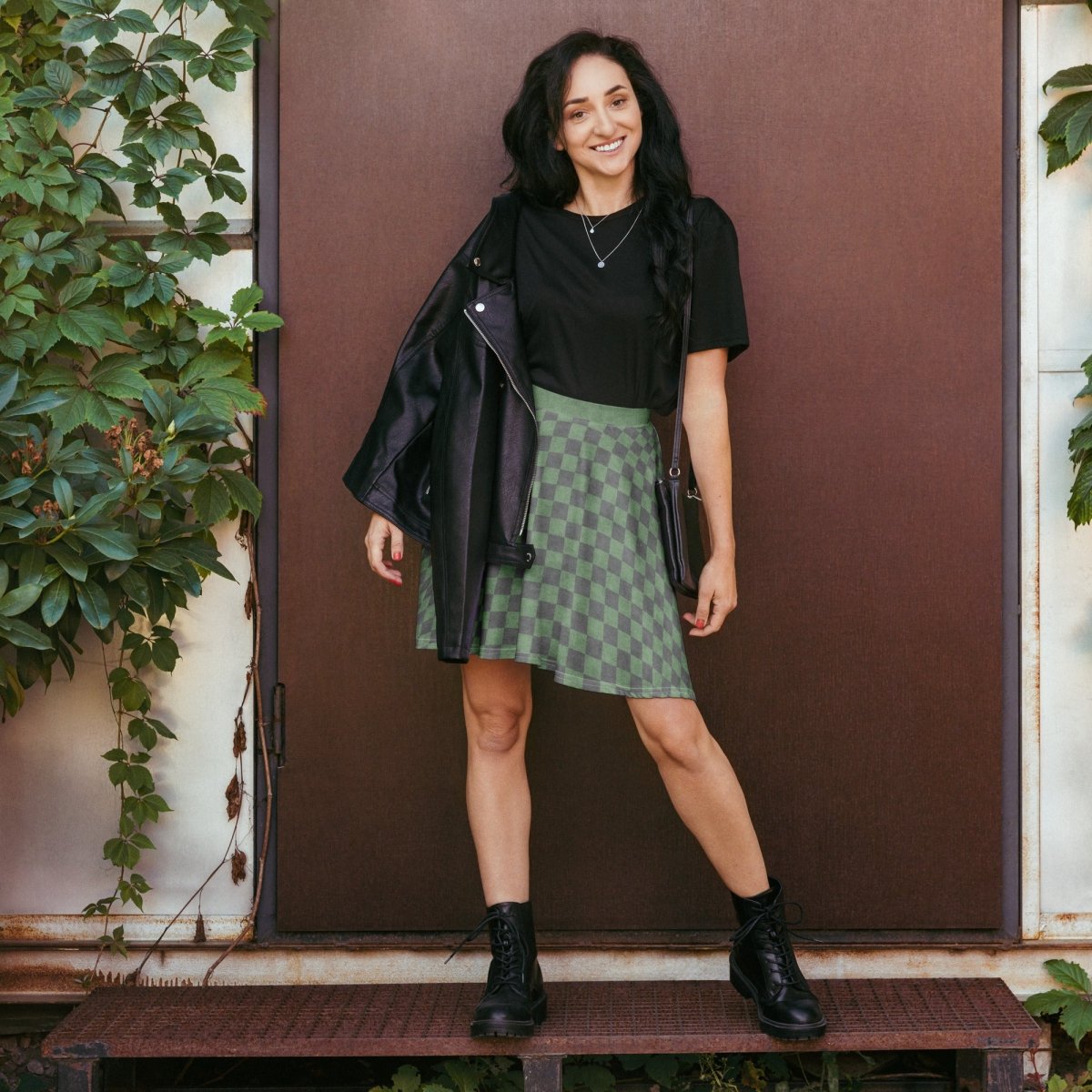 Woman standing in front of a brown door wearing a black top, green checkered skirt, and black boots.
