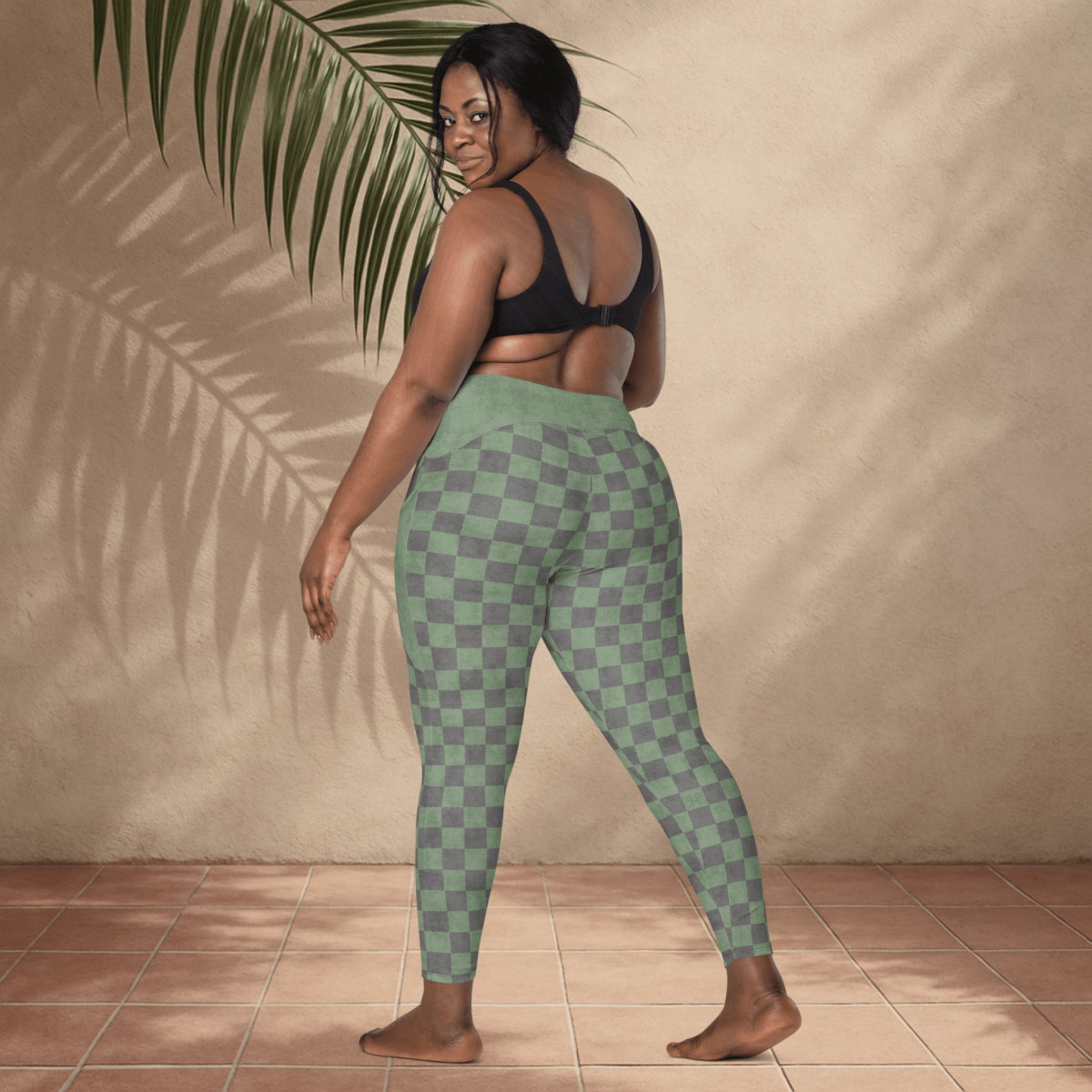 Woman wearing green checkered leggings standing against a beige wall with a plant leaf.