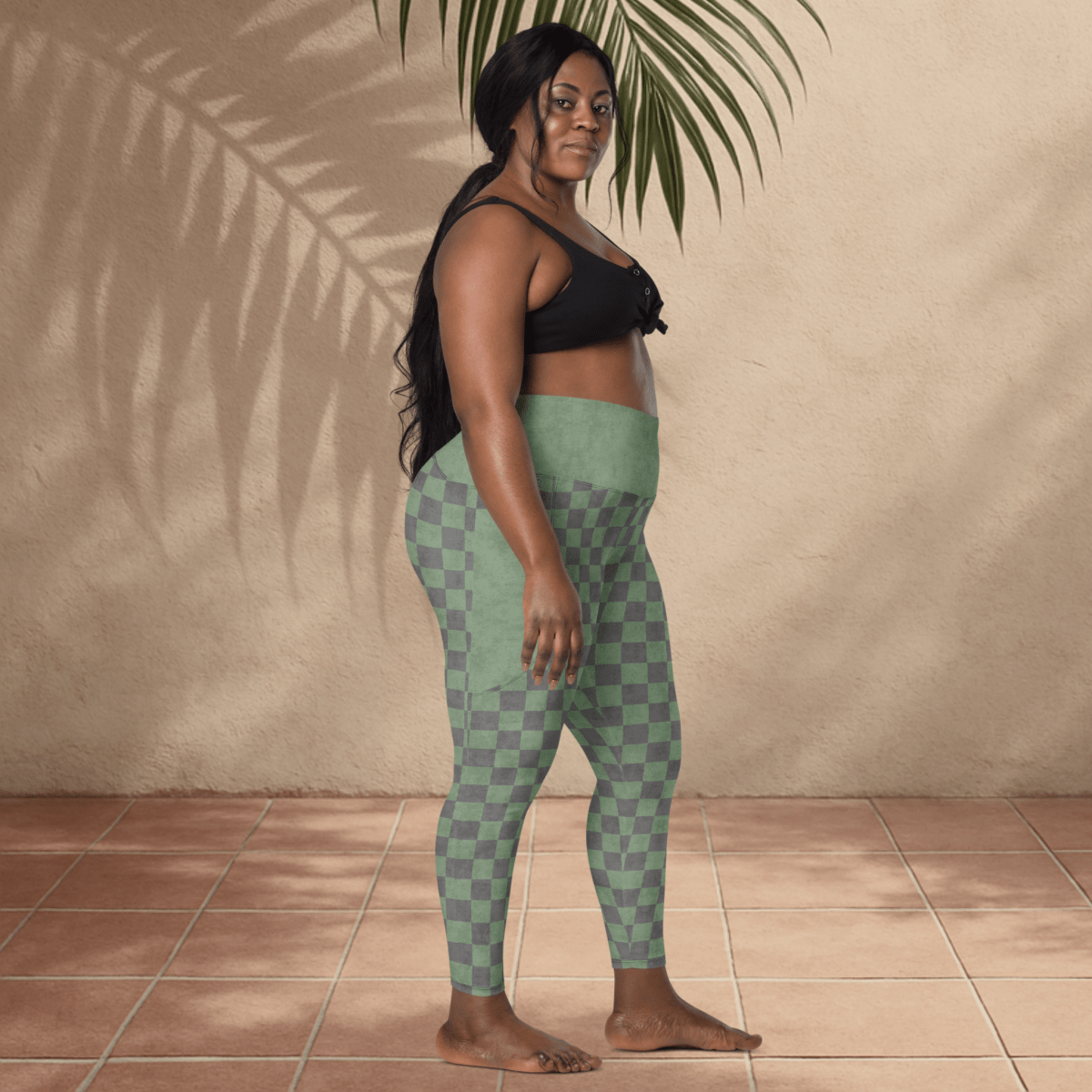 Woman wearing green checkered leggings and black top standing against a beige wall.
