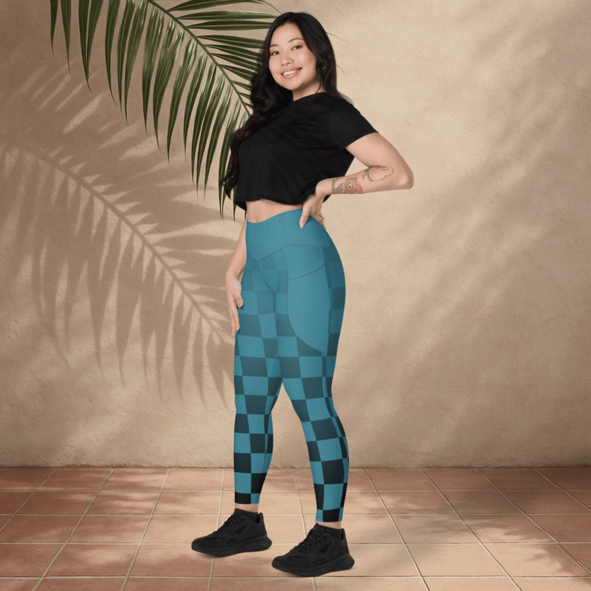 Woman wearing blue checkered leggings and black top standing against a beige wall with a plant.