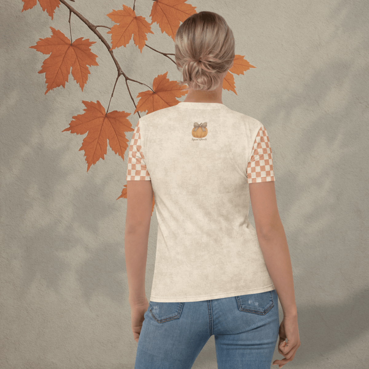 Person wearing a cream t-shirt with checkered sleeves and a pumpkin design, standing against a textured beige background with autumn leaves.