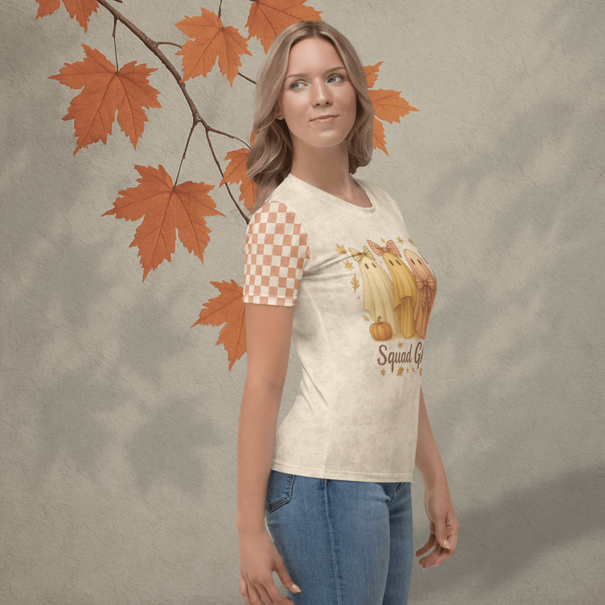 Woman wearing a t-shirt with a graphic design of pumpkins, ghosts and checkered pattern, standing against a textured background with autumn leaves.