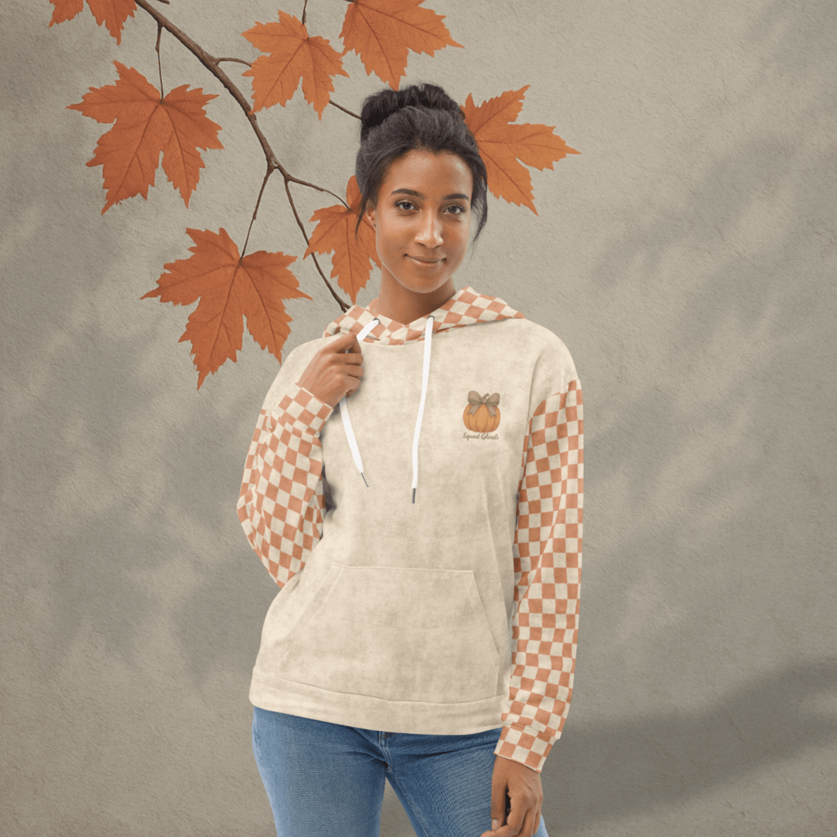 Woman wearing a beige hoodie with checkered sleeves standing next to autumn leaves.