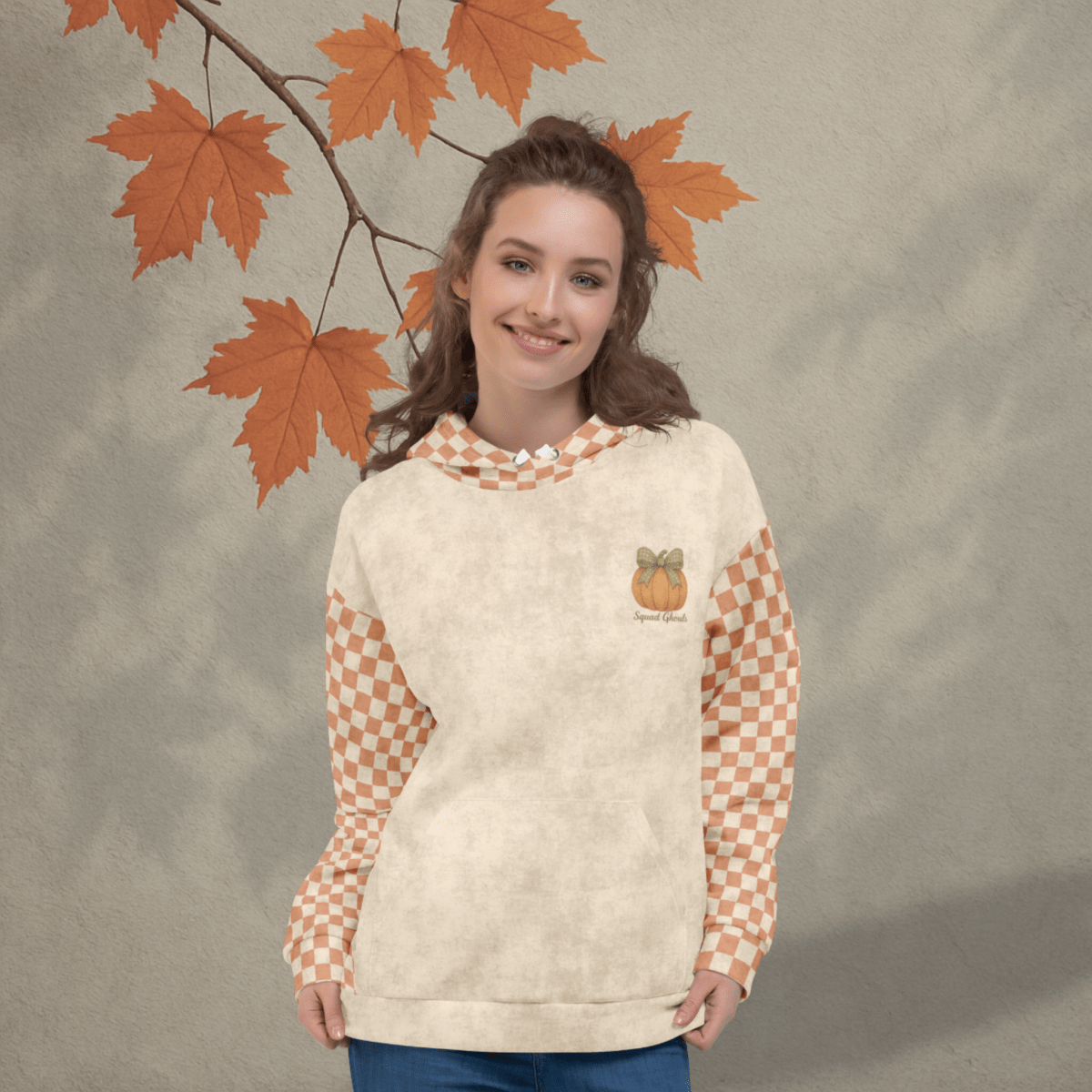 Woman wearing a cream-colored sweatshirt with checkered sleeves and a pumpkin design, standing against a neutral background with autumn leaves.