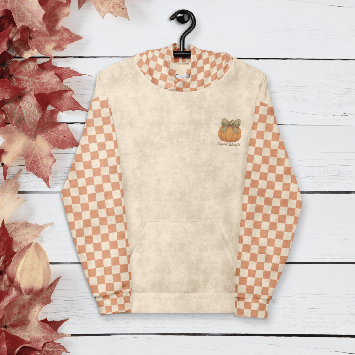 Beige hoodie with checkered sleeves on a hanger against a wooden background with autumn leaves.