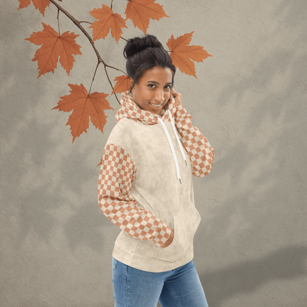 Woman wearing a beige hoodie with checkered sleeves and blue jeans, standing against a neutral background with autumn leaves.