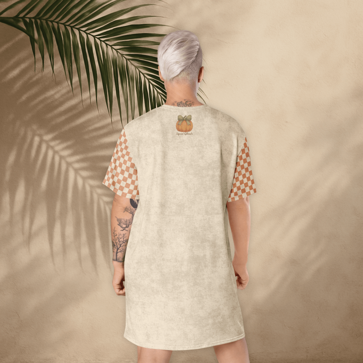 Person wearing a beige dress with a pumpkin design, standing against a beige wall with a palm leaf.