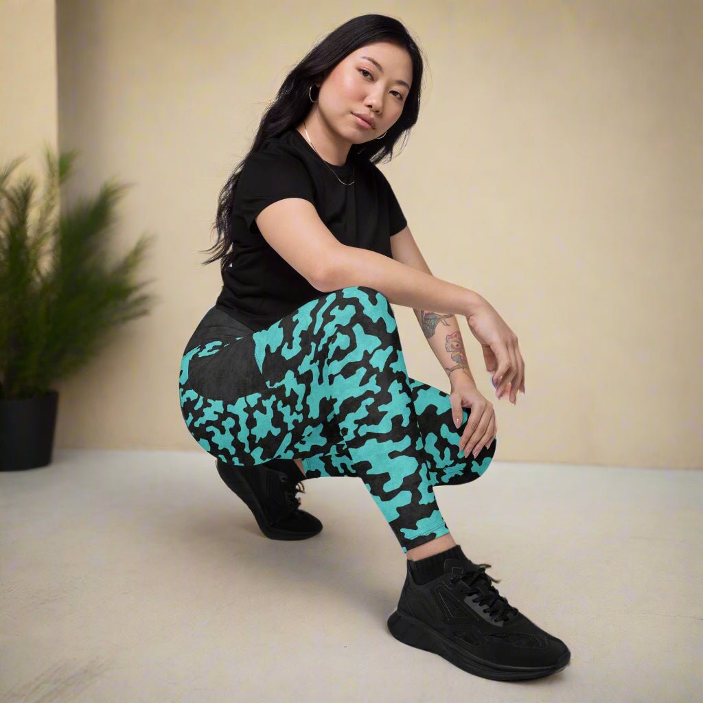 Person wearing black t-shirt and teal camouflage leggings posing against a beige wall.