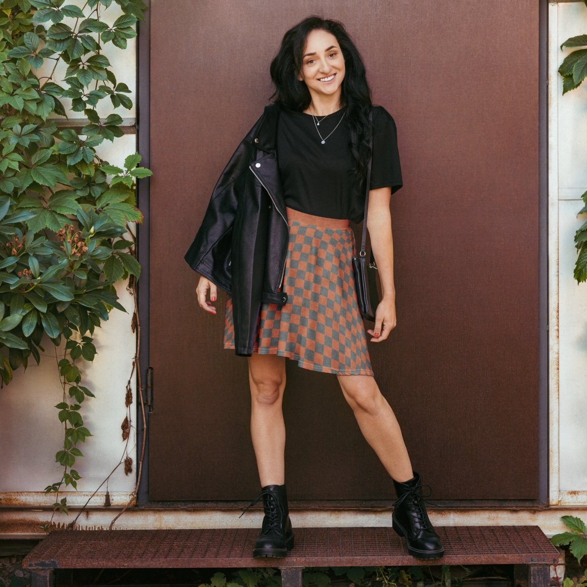Woman standing in front of a brown door wearing a black jacket, black top, and checkered skirt.