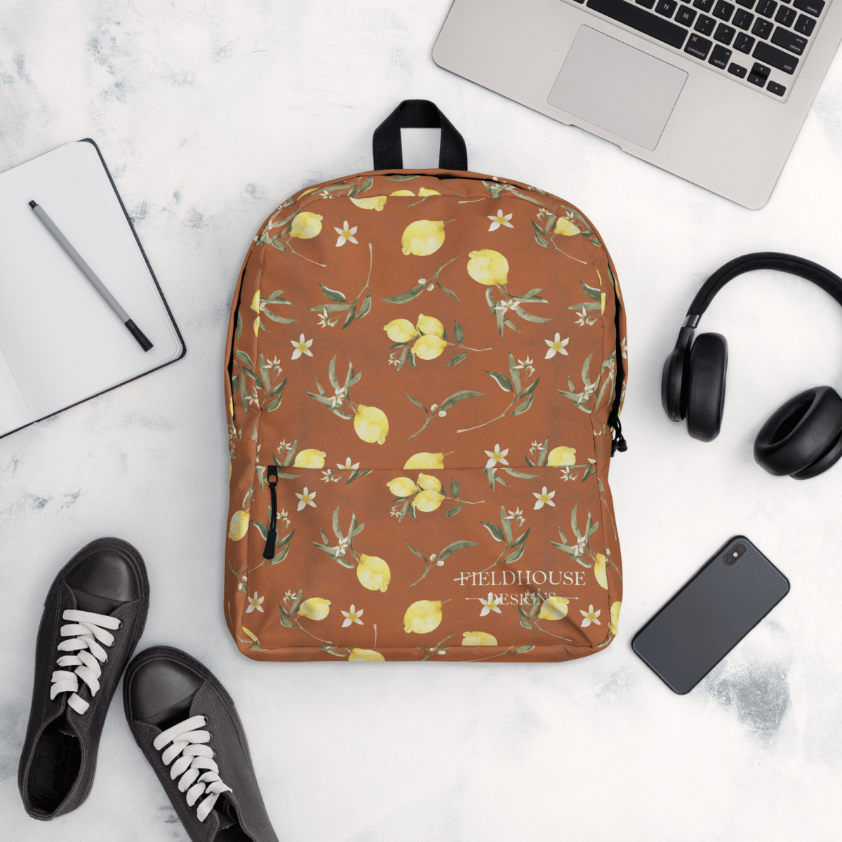 rust backpack with lemon pattern on a white surface with laptop, phone, and shoes.