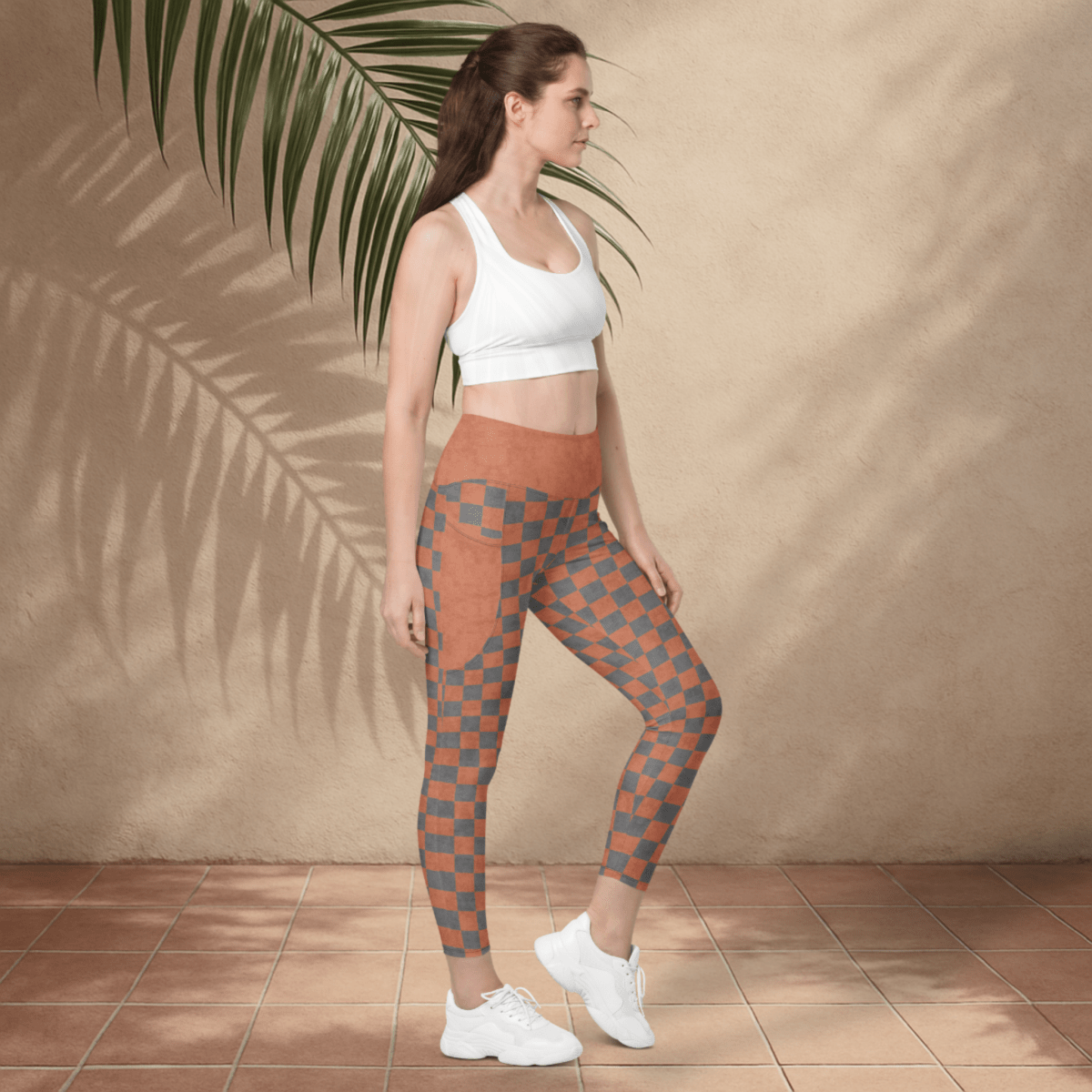 Woman wearing a white sports bra and patterned leggings standing against a beige wall with a palm leaf.