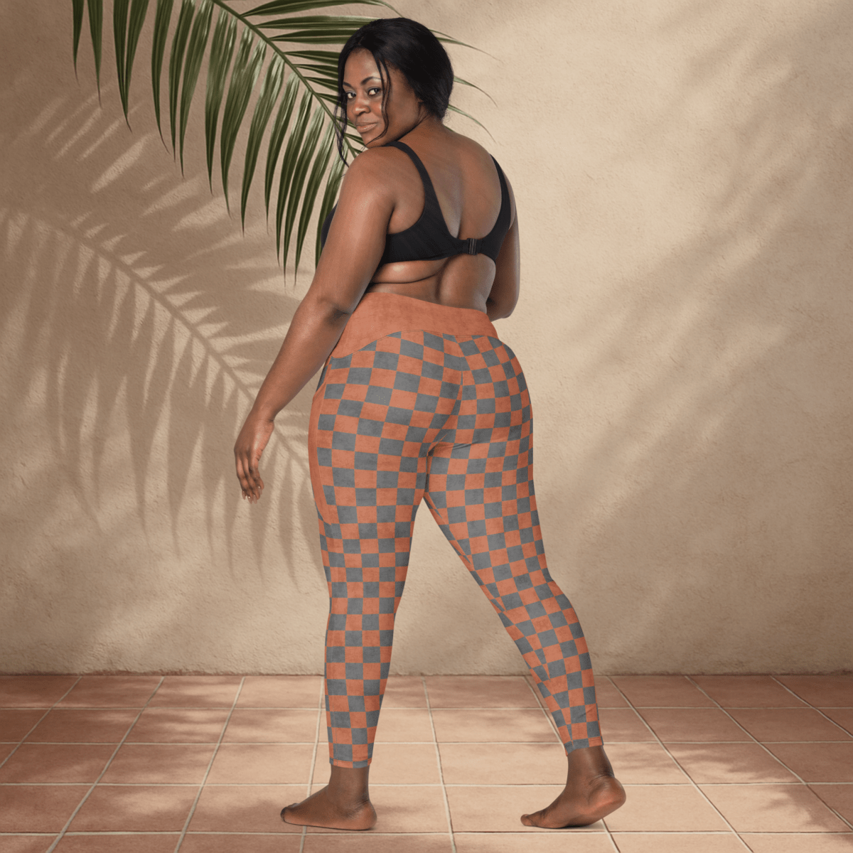 Woman wearing checkered leggings standing against a beige wall with a plant leaf.