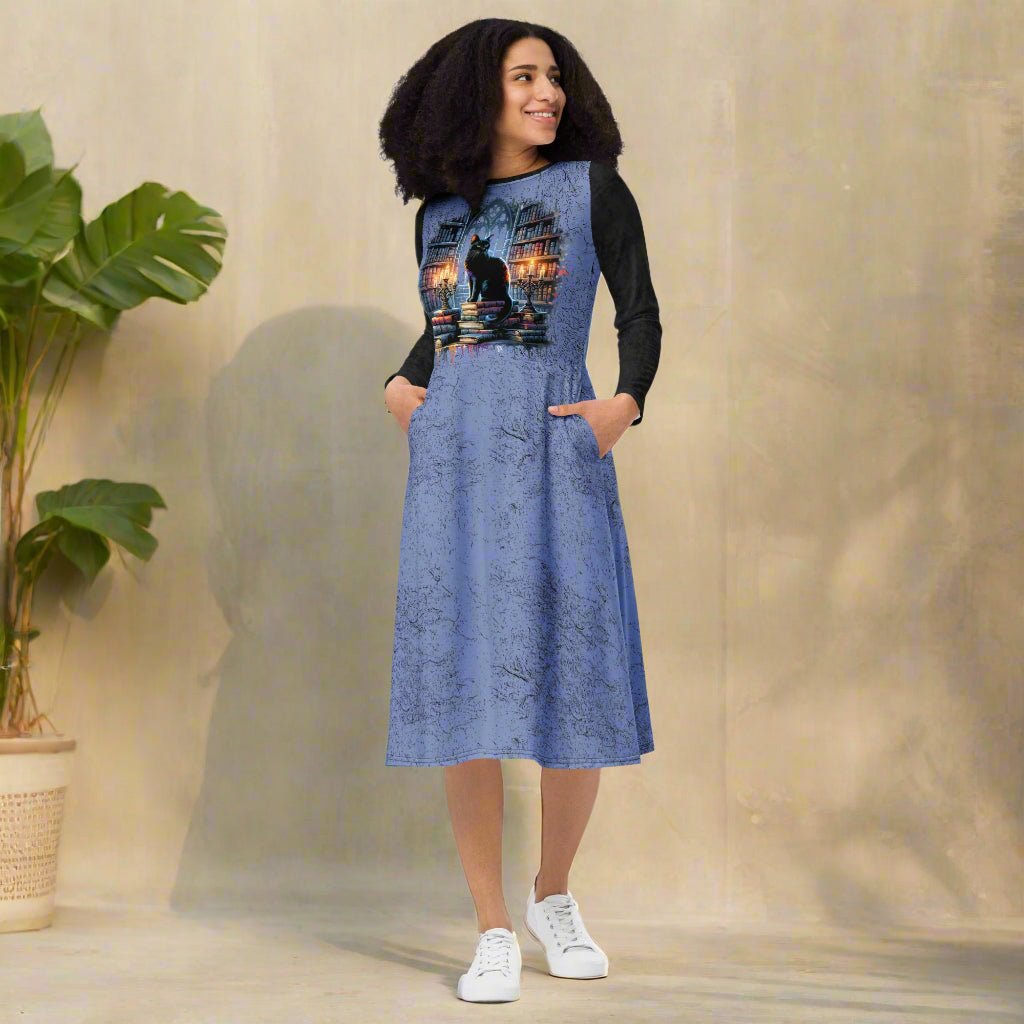 Woman wearing a blue dress with a black cat design, standing indoors next to a plant.
