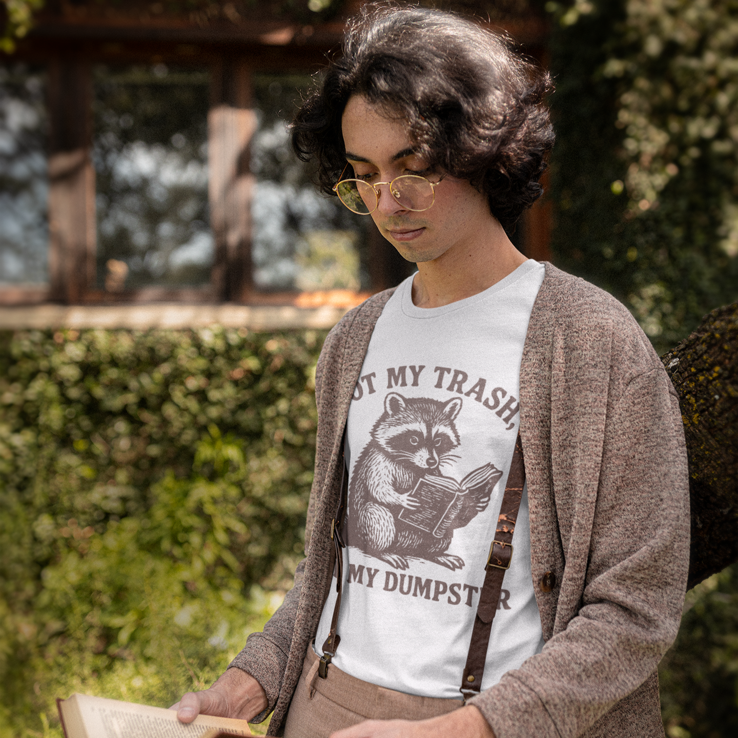 Person wearing a t-shirt with a raccoon graphic and suspenders, holding a book outdoors.