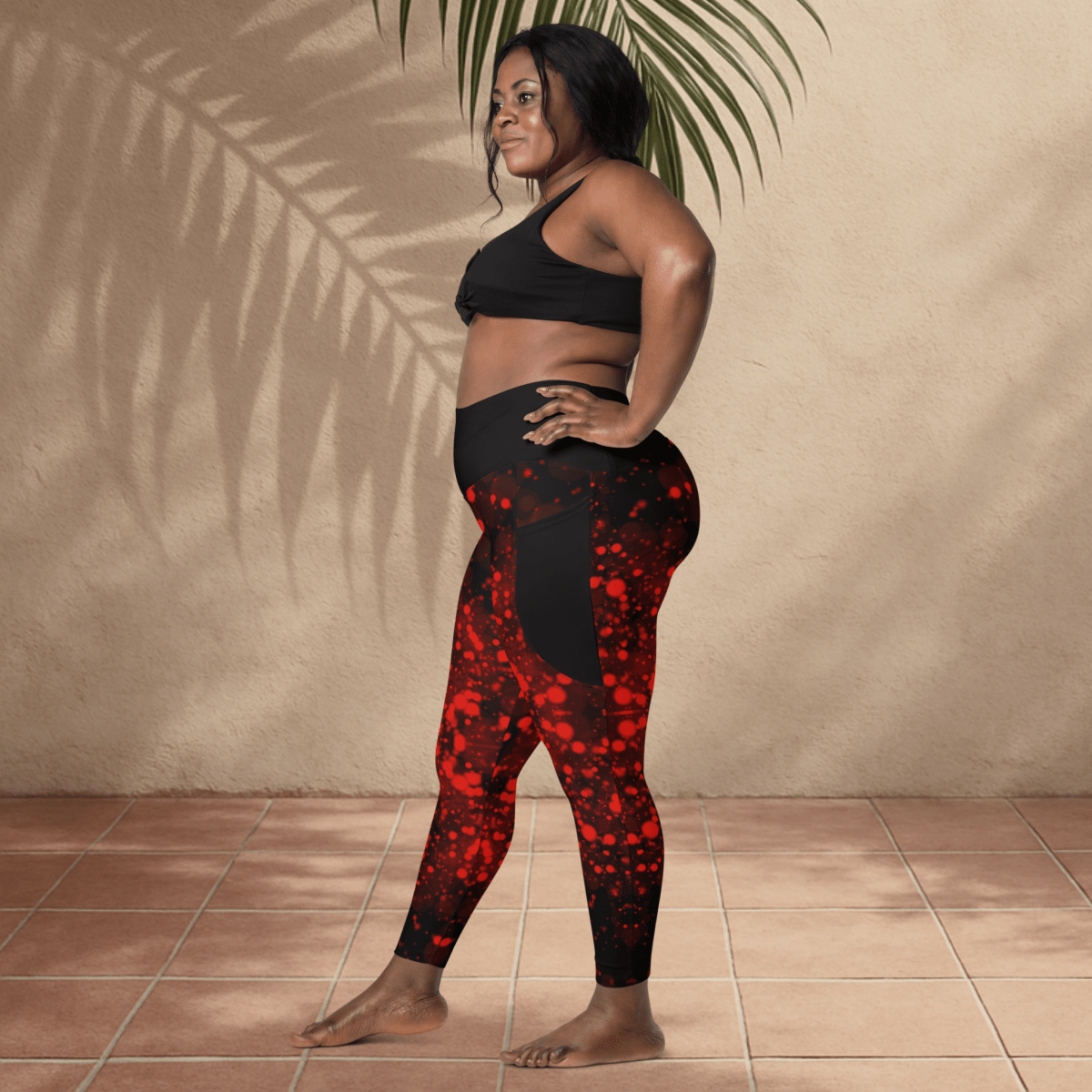 Woman wearing black sports bra and black with red patterned leggings standing against a beige wall.