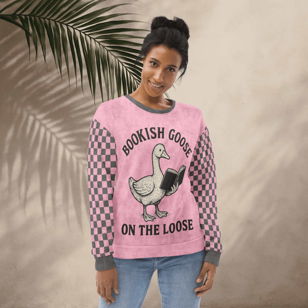 Woman wearing a pink sweater with a goose graphic and text, standing against a beige wall with a leafy branch.