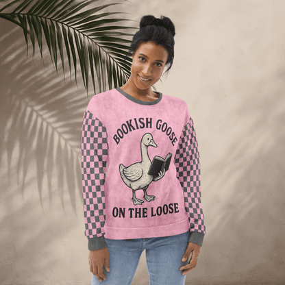 Woman wearing a pink sweater with a goose graphic and text, standing against a beige wall with a leafy branch.