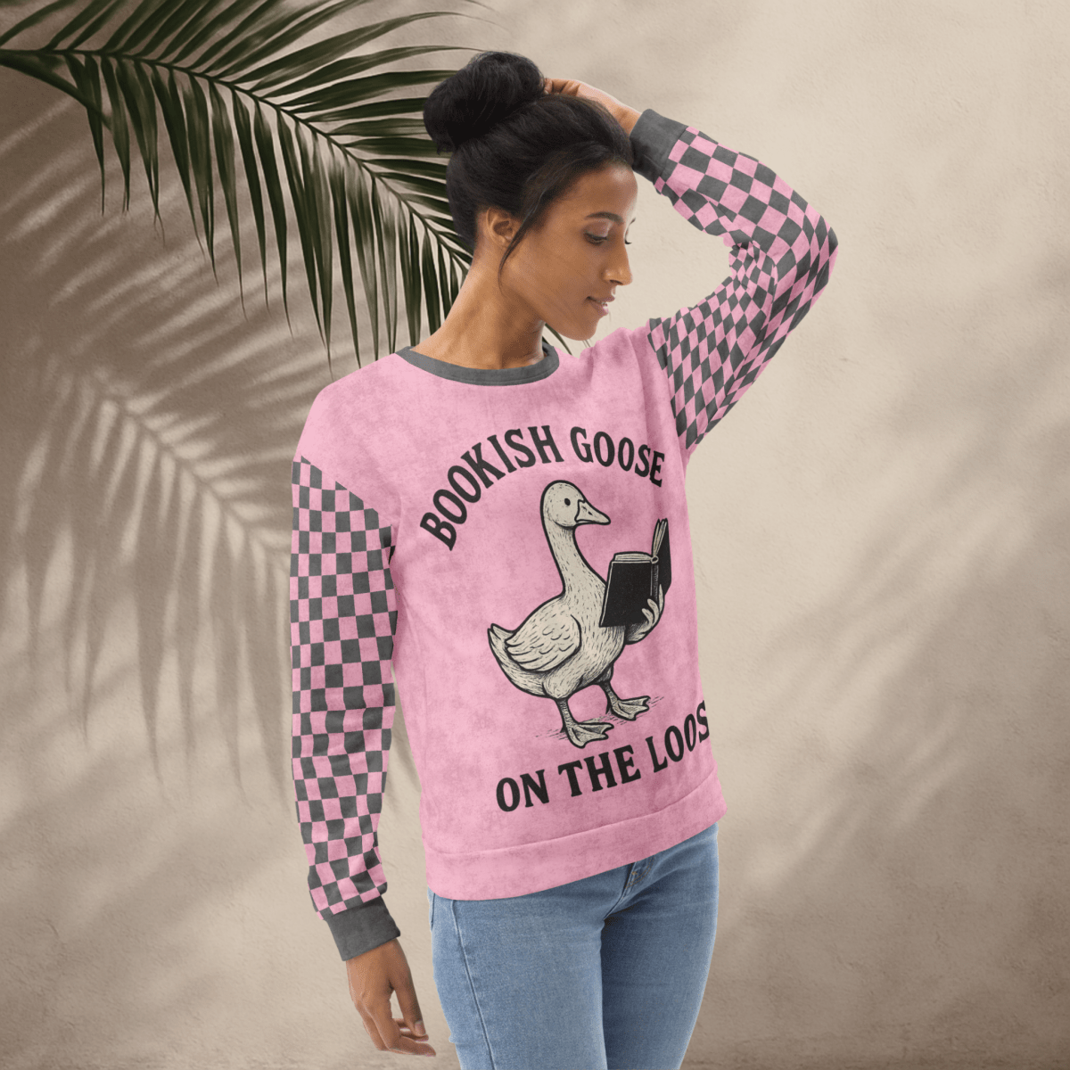 Woman wearing a pink sweater with a goose graphic and text, standing against a beige wall with a leafy branch.
