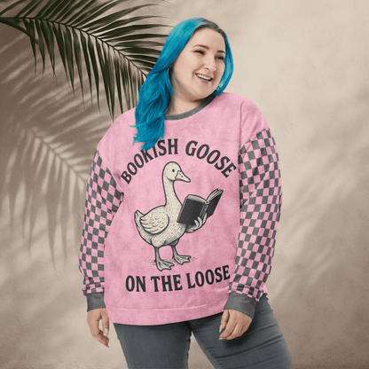 Person wearing a pink sweater with a goose graphic and text, standing against a beige wall with a leafy branch.