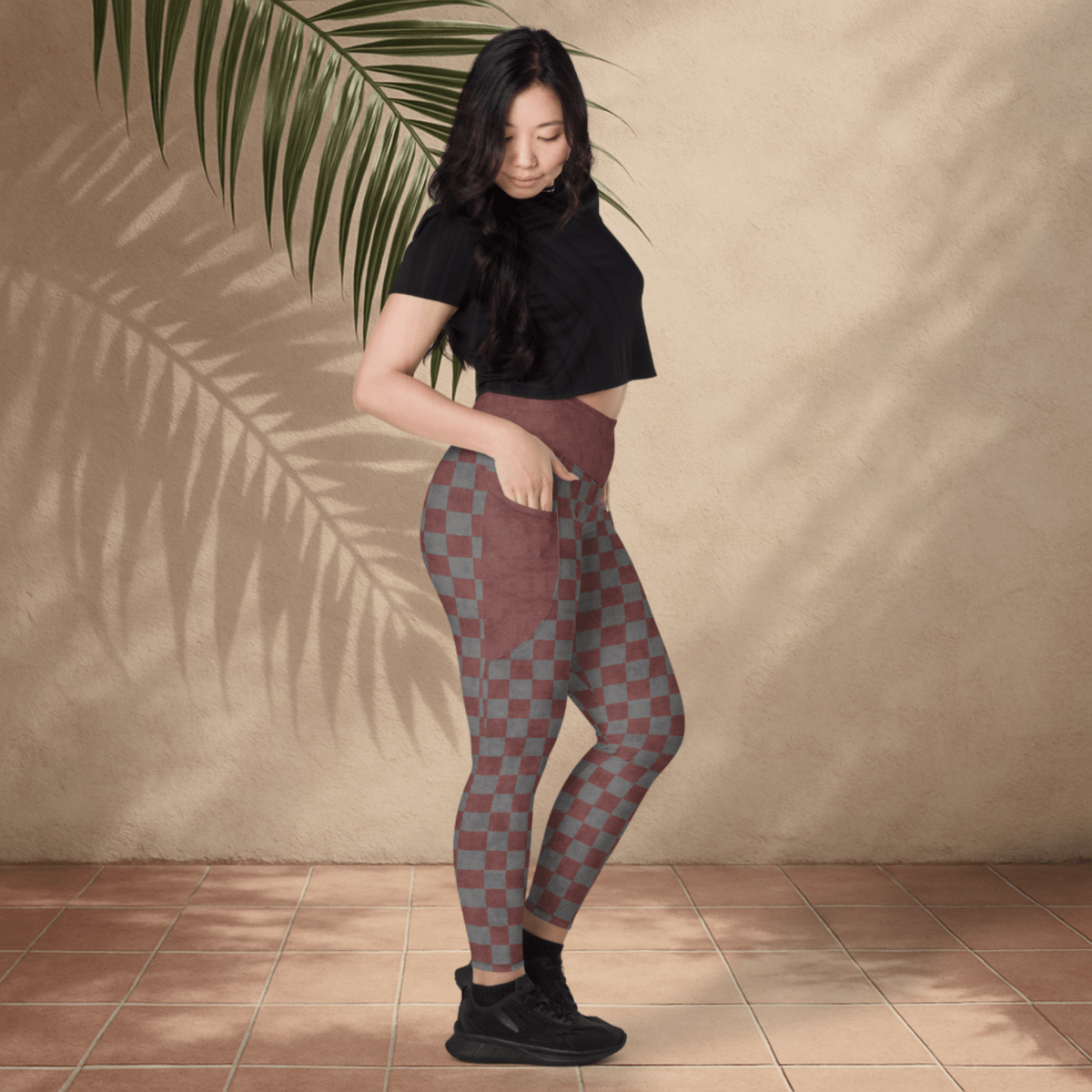 Woman wearing checkered leggings and a black top against a beige wall with a plant leaf.