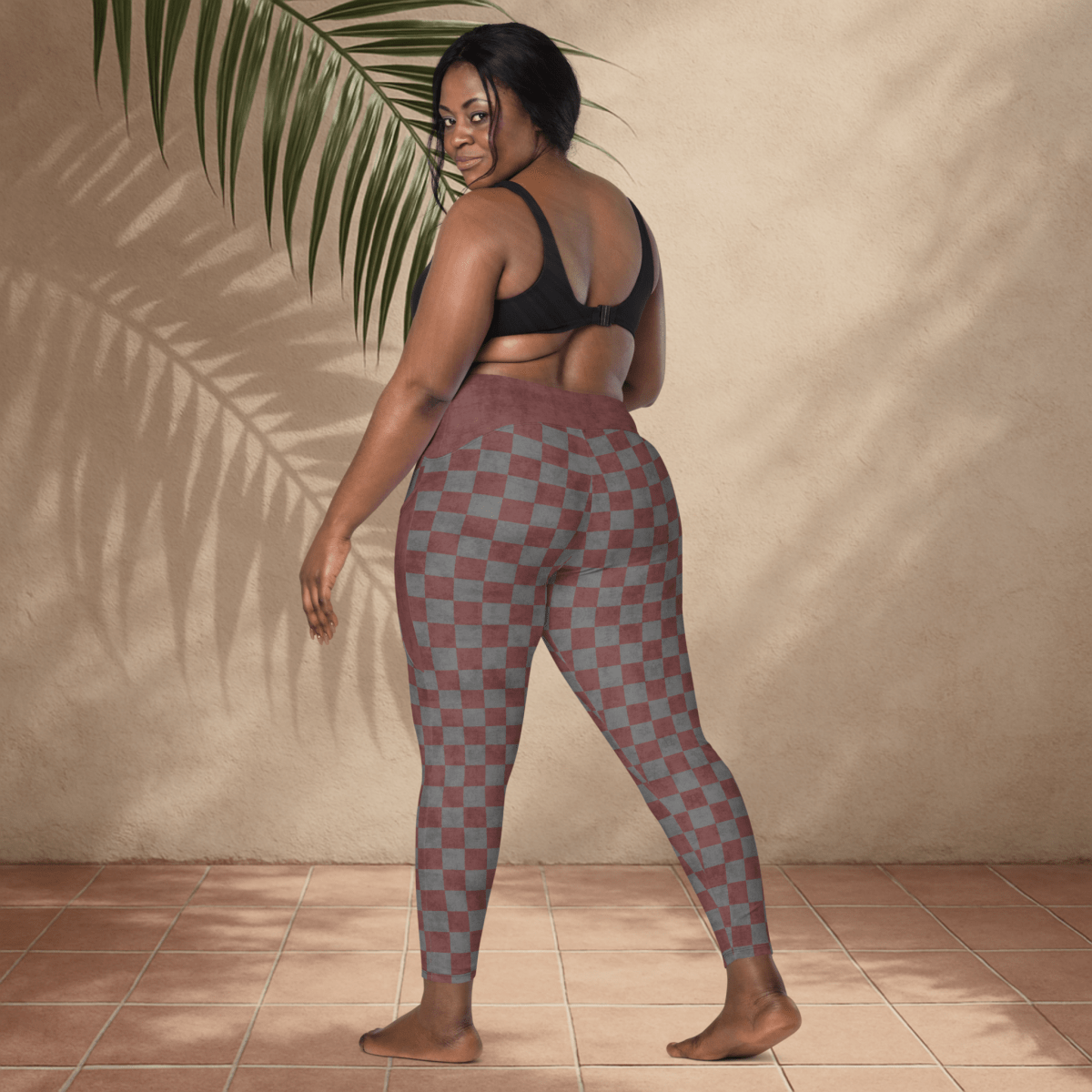 Woman wearing checkered leggings and black sports bra against a beige wall with a plant.