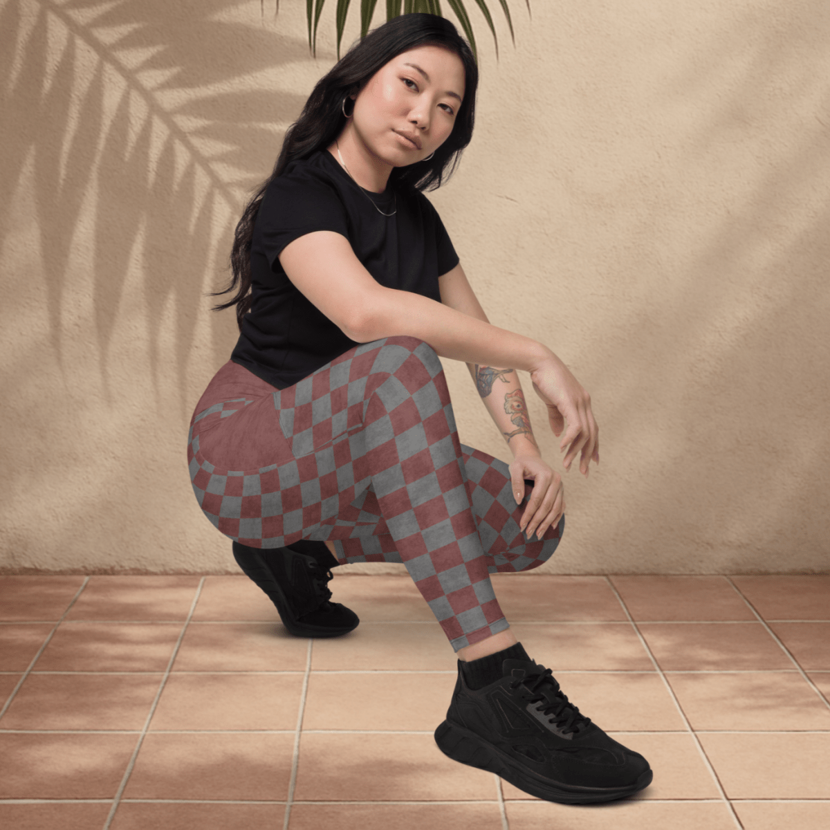 Person wearing a black t-shirt and checkered pants sitting on the floor with a leaf above them against a beige wall.