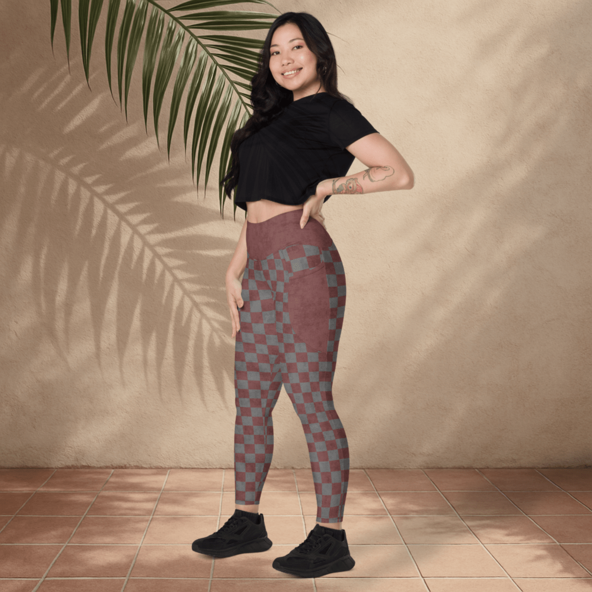 Woman wearing checkered leggings and a black top standing against a beige wall with a plant.