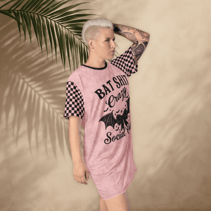 A person with platinum-blonde hair and arm tattoos poses in a pink t-shirt dress against a tan wall with palm leaf shadows. The dress features black checkerboard sleeves and a bold black graphic that reads "BAT SHIT Crazy Social Club" with a bat-winged bow and stars. The person stands with one hand on their head, tilting slightly forward.
