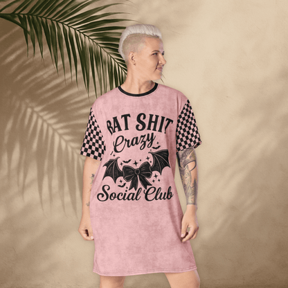A person with platinum-blonde short hair and visible arm tattoos stands in front of a tan stucco wall with palm leaf shadows, wearing a pink t-shirt dress. The dress features black checkerboard sleeves and bold black text on the front that reads "BAT SHIT Crazy Social Club," accompanied by graphic elements like a bow, bat wings, and scattered stars. The person faces forward with a slight smile and hands relaxed at their sides.