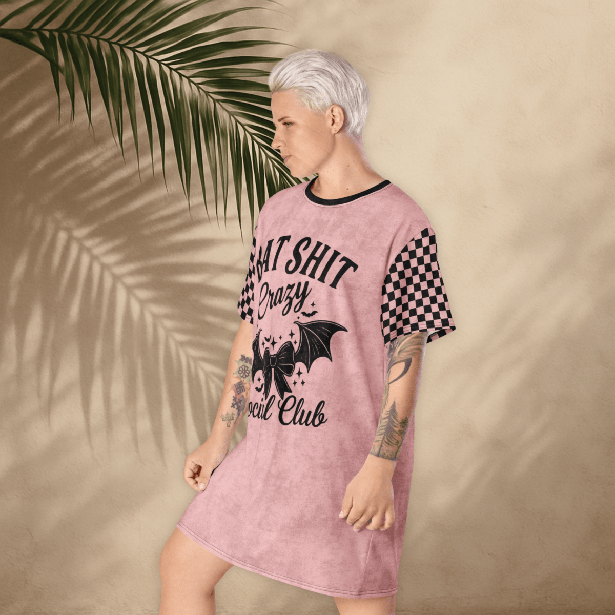 A person with platinum-blonde hair stands in a pink t-shirt dress with a moody expression and one foot slightly raised. The dress features black checkerboard sleeves and a large black front print reading "BAT SHIT Crazy Social Club" with a decorative bow and bat wings. The tan background includes soft palm shadows.