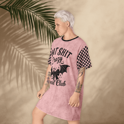 A person with platinum-blonde hair stands in a pink t-shirt dress with a moody expression and one foot slightly raised. The dress features black checkerboard sleeves and a large black front print reading "BAT SHIT Crazy Social Club" with a decorative bow and bat wings. The tan background includes soft palm shadows.