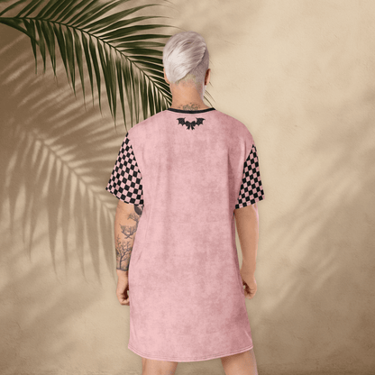 A person with platinum-blonde hair stands with their back to the camera, wearing a pink t-shirt dress. The dress has black checkerboard sleeves and a small black bat-and-bow graphic printed between the shoulder blades. The background is a light tan stucco wall with large palm leaf shadows cast across it. The person’s tattoos are visible on both arms.
