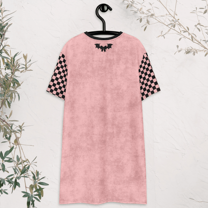 A pink t-shirt dress is shown from the back, hanging on a black hanger against a white textured wall. The dress features black checkerboard sleeves and a small black bow and bat-wing graphic centered near the neckline. Green branches frame the edges of the image.