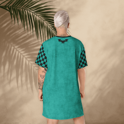 A person with platinum-blonde hair stands with their back to the camera, wearing a teal t-shirt dress. The dress has black checkerboard sleeves and a small black bat-and-bow graphic printed between the shoulder blades. The background is a light tan stucco wall with large palm leaf shadows cast across it. The person’s tattoos are visible on both arms