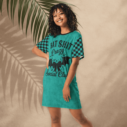 A person with medium-toned skin and curly dark hair poses smiling in a teal t-shirt dress in front of a tan stucco wall with palm leaf shadows. The dress has black checkerboard sleeves and a front design that reads "BAT SHIT Crazy Social Club" in bold black font, with a bow and bat wings below the text. 