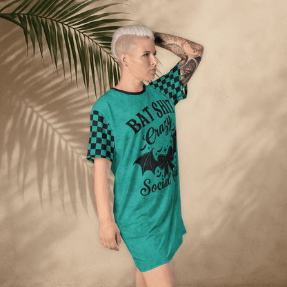 A person with platinum-blonde hair and arm tattoos poses in a teal t-shirt dress against a tan wall with palm leaf shadows. The dress features black checkerboard sleeves and a bold black graphic that reads "BAT SHIT Crazy Social Club" with a bat-winged bow and stars. The person stands with one hand on their head, tilting slightly forward.