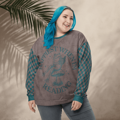 Person wearing an espresso and teal sweatshirt with a Frog reading with "Happiest When I'm Reading" , standing against a beige wall with a leafy branch.