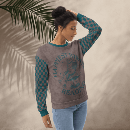 Woman wearing an espresso and teal  sweatshirt with a Frog reading with "Happiest When I'm Reading"  design and checkered sleeves against a beige background