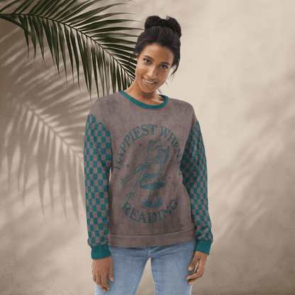 Woman wearing an espresso and teal  sweatshirt with a Frog reading with "Happiest When I'm Reading" and checkered sleeves, standing against a beige wall with a palm leaf.