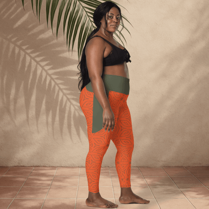 Woman wearing black sports bra and orange leggings with green accents, standing against a beige wall with a plant leaf.