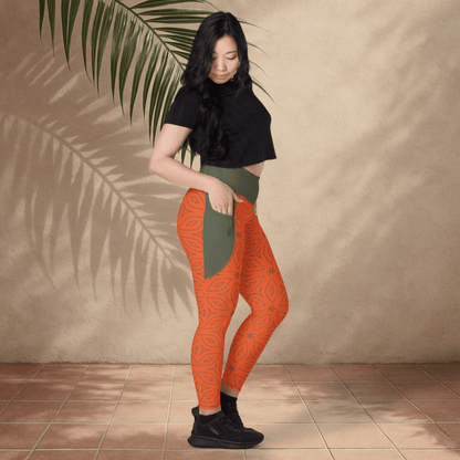 Woman wearing orange leggings with a green pattern, standing against a beige wall with a leafy branch.
