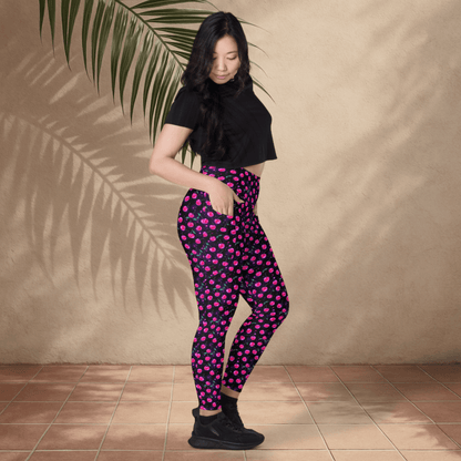 Woman wearing black crop top and pink cherry print leggings standing against a beige wall with a plant leaf.