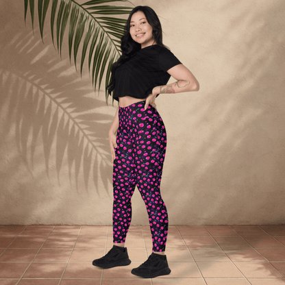 Woman wearing black crop top and pink cherry print  on black pants standing against a beige wall with a palm leaf.
