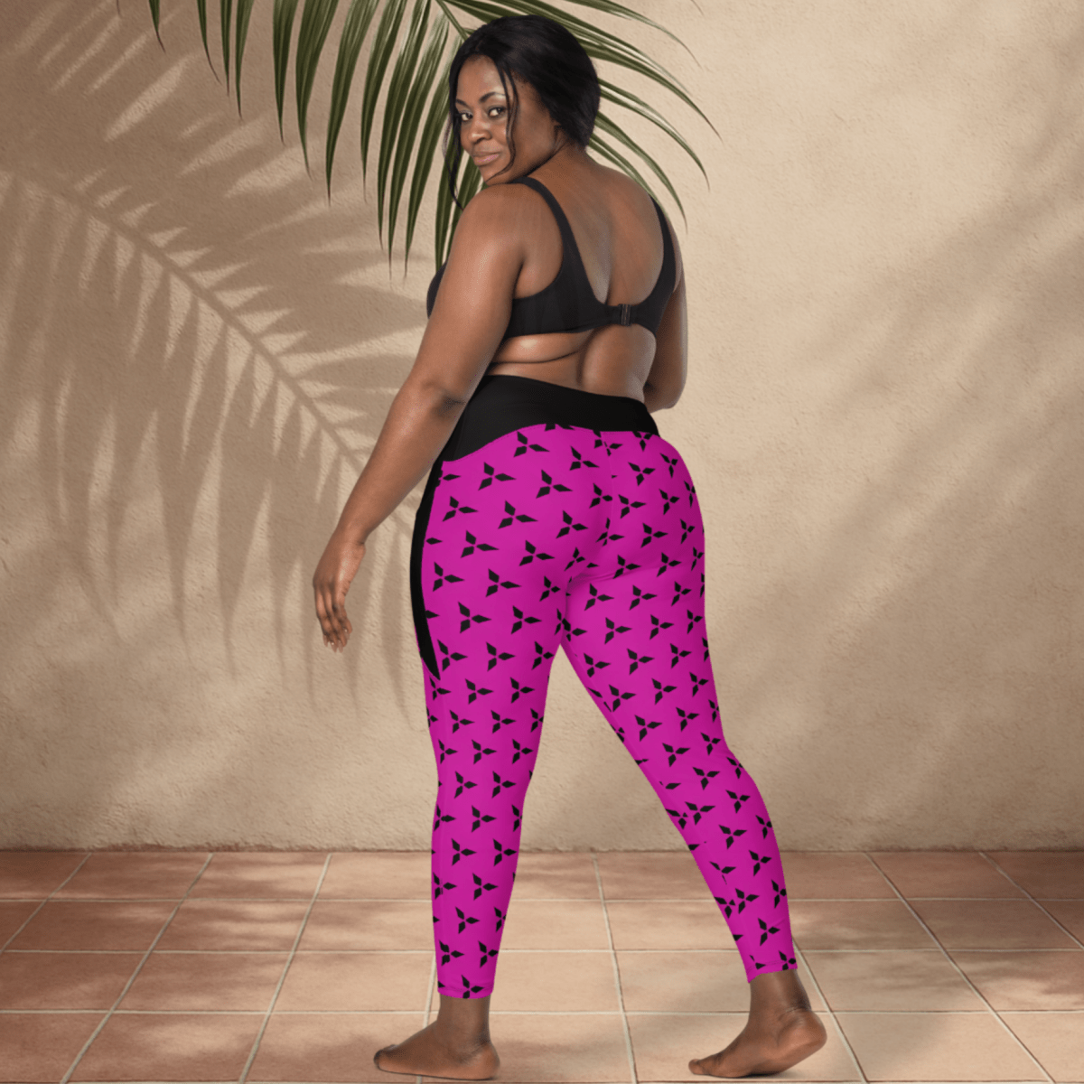 Woman wearing pink leggings with black patterns and a black sports bra against a beige wall.