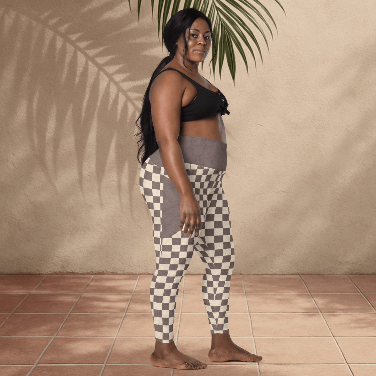 Woman wearing black sports bra and checkered leggings standing against a beige wall with a plant.