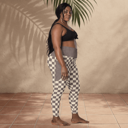 Woman wearing black sports bra and checkered leggings standing against a beige wall with a plant.