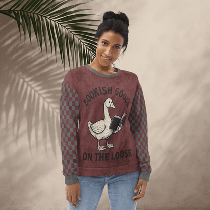 Woman wearing a maroon sweatshirt with a graphic of a goose holding a book and text 'Bookish Goose on the Loose', standing against a beige wall with a palm leaf.