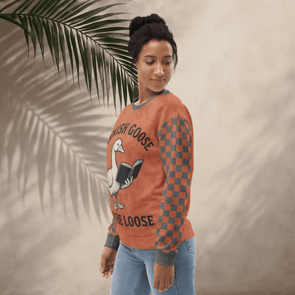 Woman wearing a terracotta checkered long-sleeve shirt with a goose reading a book graphic and text 'Bookish Goose on the Loose', standing against a beige wall with a palm leaf.
